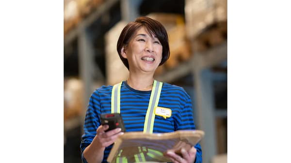 IKEA co-worker in blue striped uniform and safety vest holding a handheld device and papers in the warehouse area.