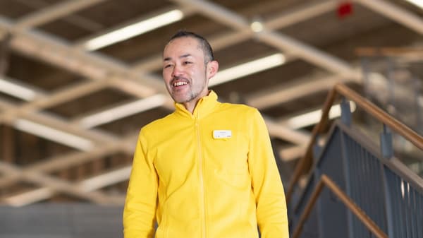 IKEA coworker in a yellow jacket with name badge walking down stairs in a bright store interior with wooden railings and ceiling beams