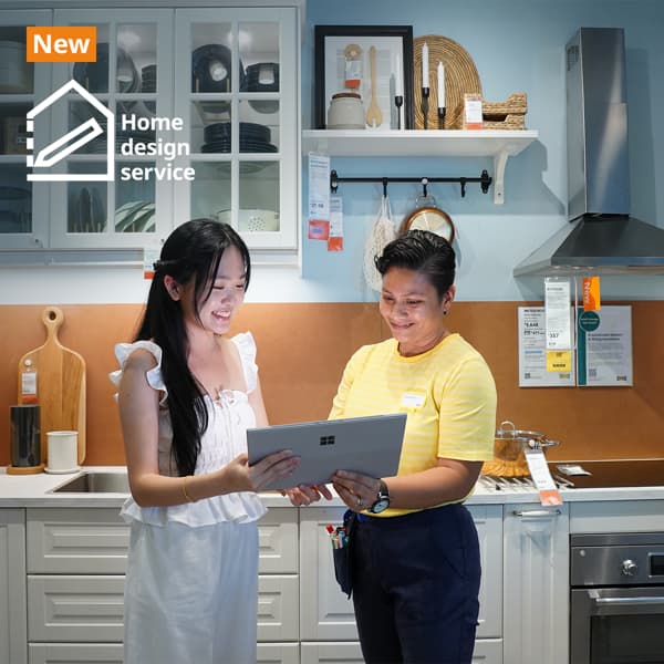 IKEA coworker with a customer in the IKEA kitchen showroom standing together holding a laptop discussing home design. They are standing infront of a METOD kitchen showcase with kitchen cabinets in white fronts and glass doors showing utensils and plates neatly displayed inside. Text on image at the top left hand corner of the image read 
