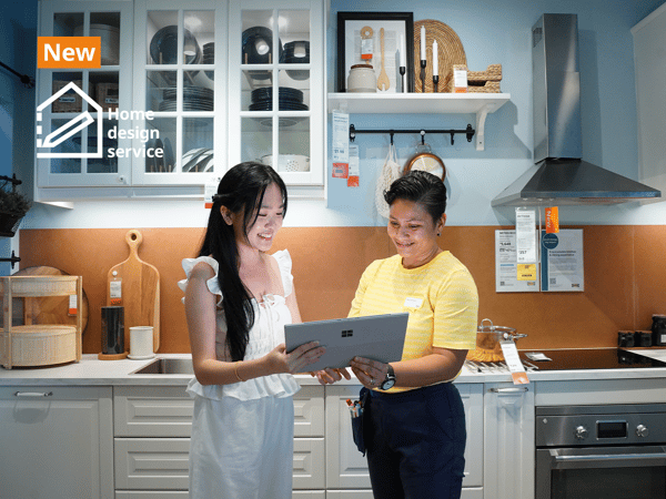 IKEA coworker with a customer in the IKEA kitchen showroom standing together holding a laptop discussing home design. They are standing infront of a METOD kitchen showcase with kitchen cabinets in white fronts and glass doors showing utensils and plates neatly displayed inside. Text on image at the top left hand corner of the image read 