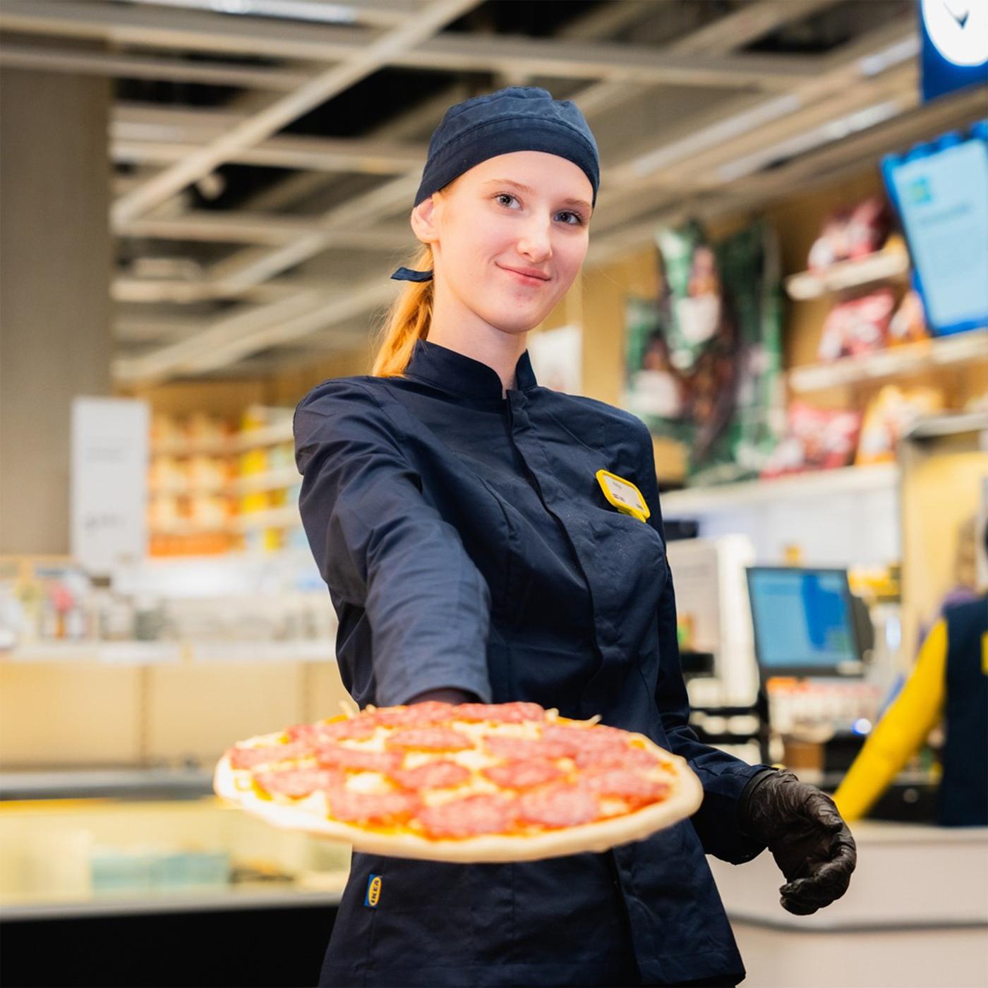 IKEA darbiniece tumši zilā uniformā pasniedz salami picu kamerā, fonā veikala iekārtojums.
