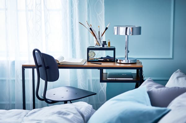 IKEA FJÄLLBO Mesa p/portátil num quarto azul, com uma cadeira preta em estilo industrial. Em cima da secretária um candeeiro de mesa STOCKHOLM, pincéis, um livro e em baixo, arrumado um computador portátil.