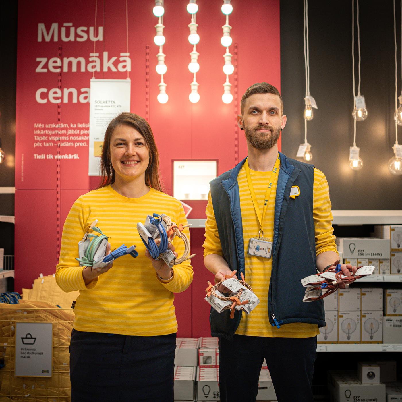 IKEA formas tērpā ģērbta eksperte un eksperts pie apgaismojuma stenda veikalā, rokās turot dažādus vadus un lādētājus.