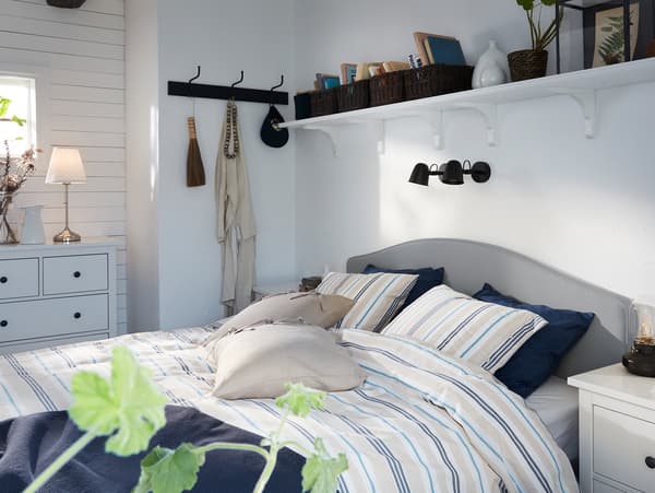 IKEA HAUGA beige upholstered bed with striped quilt cover and pillowcases and two black wall lamps mounted on the wall above.