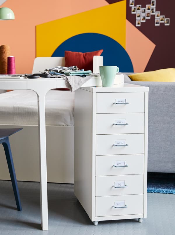 IKEA HELMER Bloco de gavetas c/rodízios encostado a uma secretária com uma caneca e um livro em cima