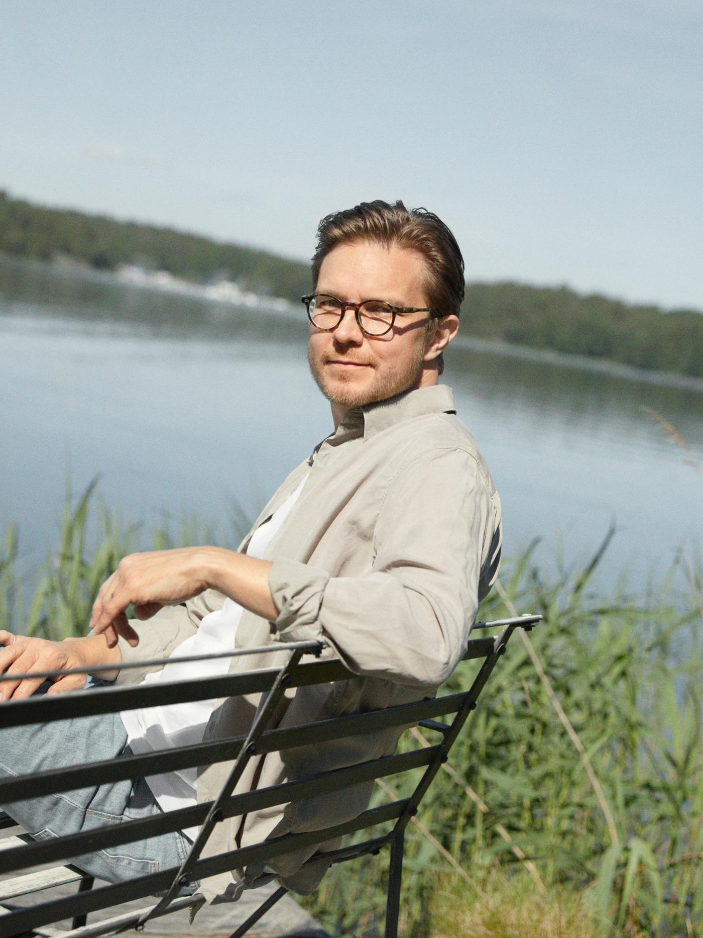IKEA 디자이너 Henrik Preutz가 밝고 화창한 날 호숫가 옆에서 벤치에 한 팔을 올려둔 채 앉아 있는 모습.