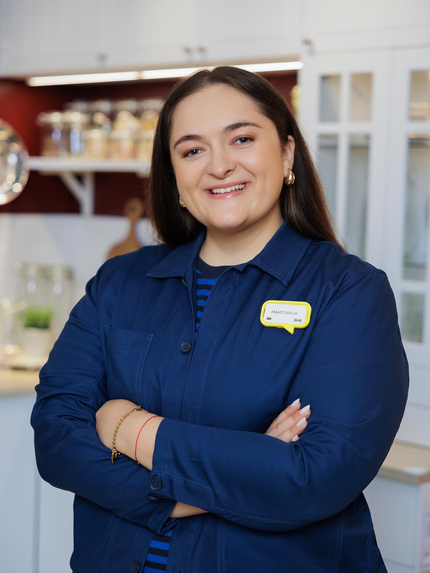 IKEA interior designer in a blue uniform stands with arms crossed, smiling confidently. A name tag is visible on their uniform.