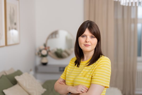 IKEA interior designer wearing a yellow striped top standing in a bedroom with her arms crossed, with a mirror and bed visible in the background.