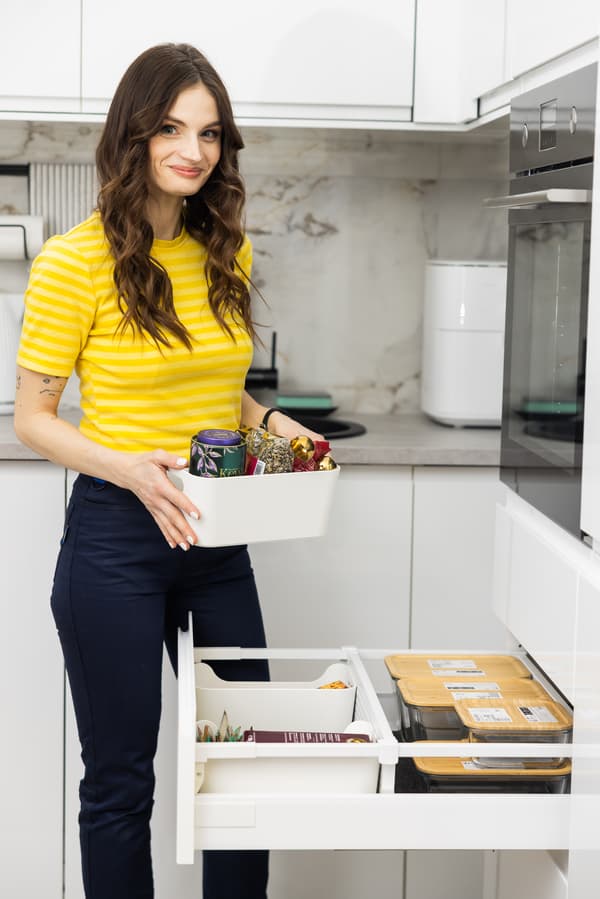 IKEA interjera dizainere stāv modernā baltā virtuvē un atver atvilktni ar organizētiem traukiem un piederumiem.