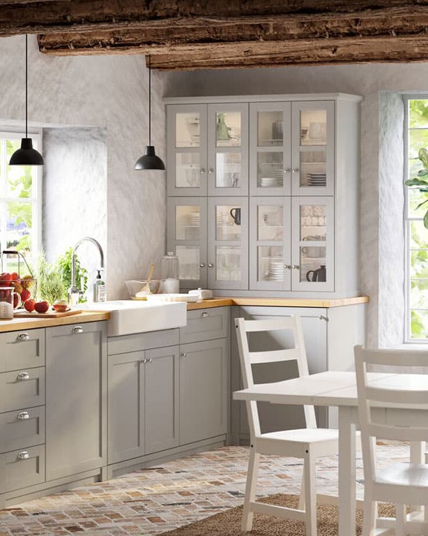 IKEA Küchenlösung im Brithischem Stile, zu sehen ist eine mittel große IKEA Küche mit Holzerner Arbeitsplatte mit einem weißen Waschbecken und grauen Unterschränken und einer passenen Vitrine. Rechts davon ein passender Esstisch mit weißen Stühlen.