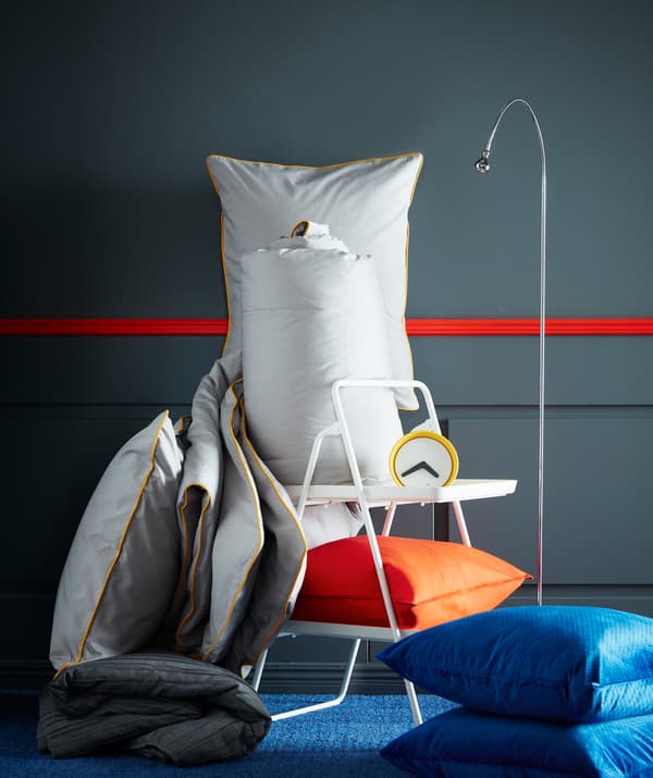 IKEA KUNGSBLOMMA siva navlaka jorgana i jastučnica sa žutim ivicama, budilnik, lampa za čitanje i narandžasti i plavi jastuci.