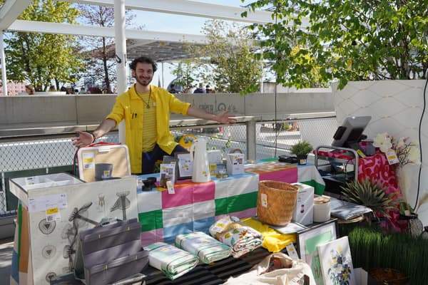 IKEA-Mitarbeiter im gelben Shirt begrüßt freundlich Besucher am nachhaltigen Second-Hand-Stand mit gebrauchten IKEA-Produkten, Wohnaccessoires und Pflanzen auf einer sonnigen Dachterrasse.