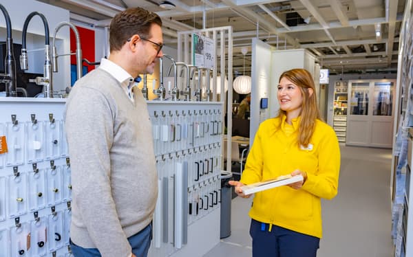  IKEA-Mitarbeiterin im gelben Outfit berät einen Kunden vor einer Wand mit Wasserhähnen und Küchen- bzw. Badgriffen. Beide lächeln und wirken im Gespräch vertieft.