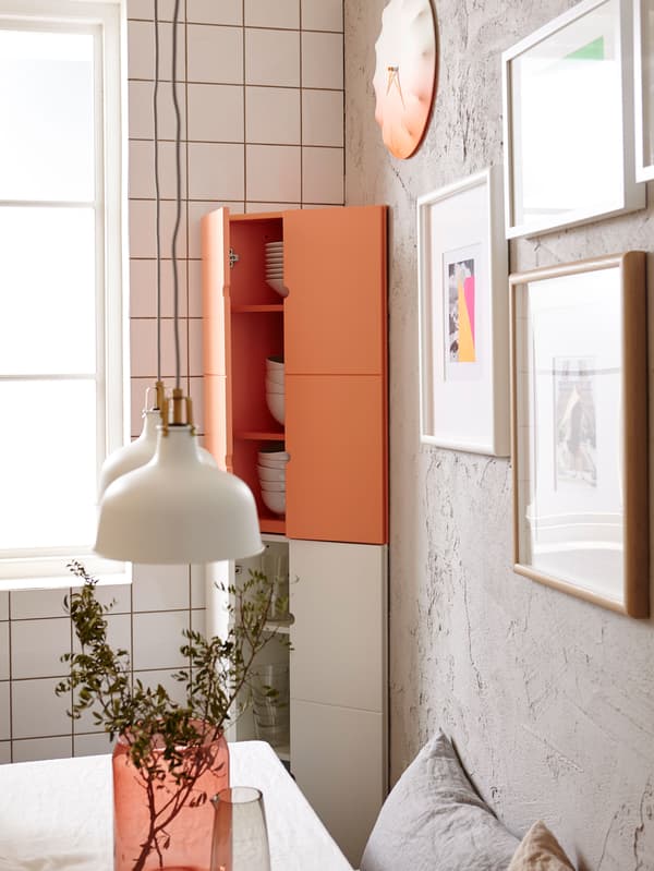 IKEA PS corner cabinets in white and orange, in a small kitchen, with pictures and a clock on the wall.