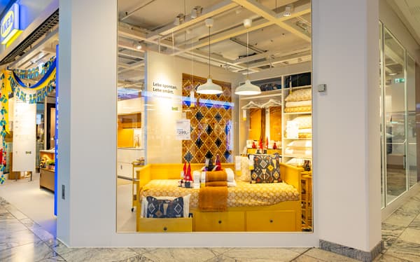 IKEA-Schaufenster mit gelbem Tagesbett, dekoriert mit bunten Kissen und schwedischen Flaggen. Im Hintergrund eine Fliesendekoration und hängende Lampen. Slogan an der Wand: „Lebe spontan. Lebe smart.“