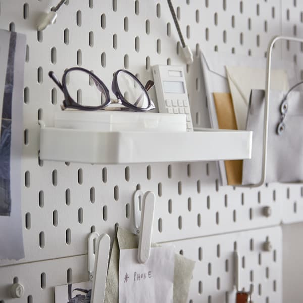 IKEA SKÅDIS Prateleira encaixada num placar com uns óculos pretos arrumados. Ao lado vê-se uma calculadora e alguns papéis arrumados.