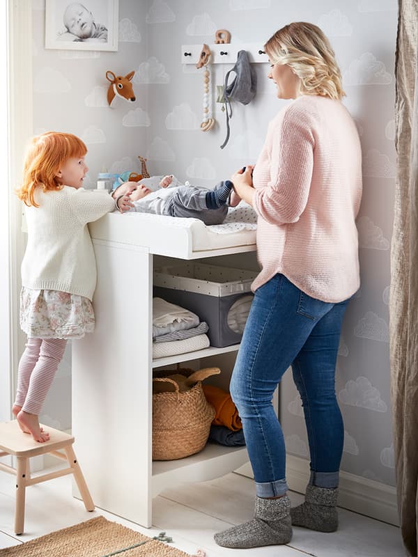 IKEA SMÅGÖRA changing table in a room with floral wallpaper, wardrobe, armchair, mobile and baby nursing items.