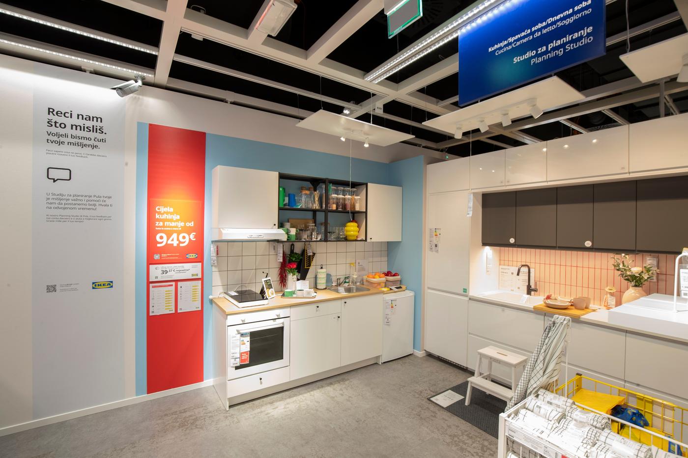 IKEA Studio za planiranje s dvije izložbene kuhinje – bijela s drvenom pločom i crvenim promidžbenim panoem s cijenom te siva s ružičastim pločicama.
