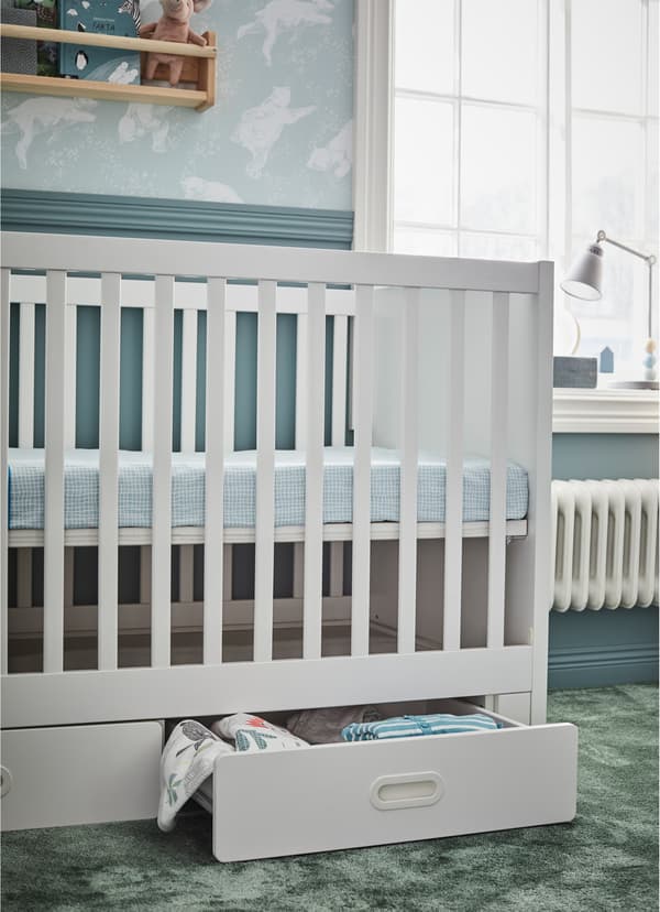 IKEA STUVA FRITIDS white cot in the high position. Lower white shelf is ajar holding baby blankets and bedsheets.