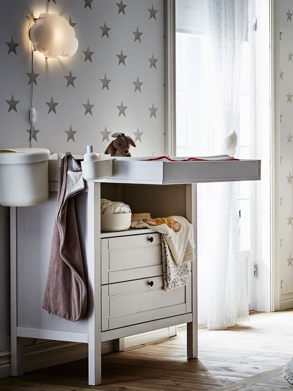 IKEA SUNDVIK changing table in white with baby clothes and baby linens, crib and small side table.