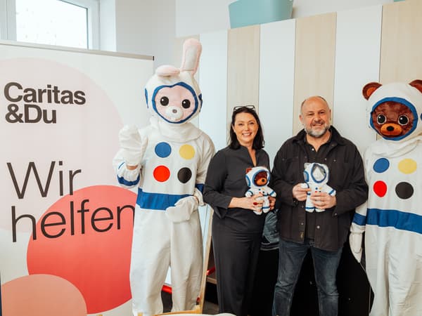IKEA und Caritas Mitarbeitende posieren mit zwei verkleideten Maskottchen in Raumanzügen bei einer sozialen Aktion; Plakat mit Aufschrift ‚Caritas &Du – Wir helfen‘ im Hintergrund