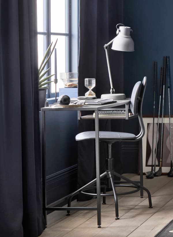 IKEA VITTSJÖ Mesa p/portátil encostada a uma janela, com uma cadeira preta em estilo industrial. Em cima da secretária caixas, alguns papéis e um candeeiro de secretária.