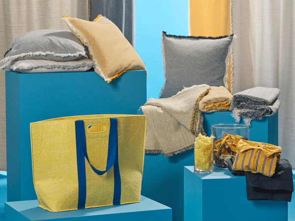 IKEA Wohnaccessoires in sommerlichen Farben auf blauen Podesten präsentiert, darunter gelbe Tragetasche, Kissen, Decken, Gläser und Heimtextilien – ideal für gemütliche und farbenfrohe Einrichtungsideen im Frühling und Sommer.