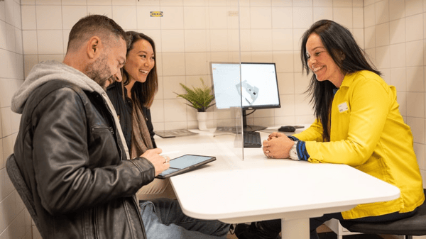 Ikea zaposlenik razgovara s parom u prostoriji iznad bijelog stola, a par se smiješi i gleda digitalni tablet.