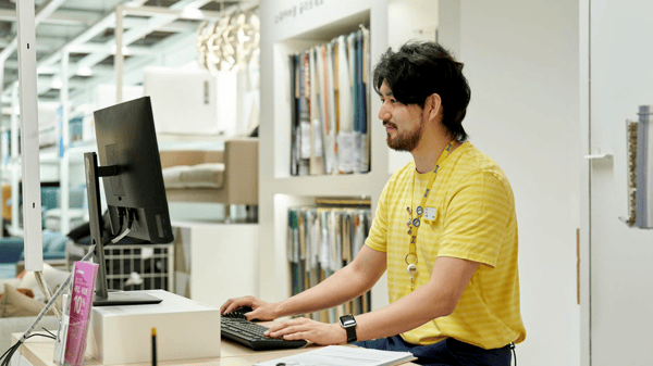 イケア店舗の売場カウンターで、コワーカーがデスクトップPCを使いながらお客さま対応やサービス業務を行っている様子。