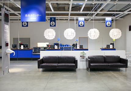 イケア店舗の返品カウンターエリアで、青い案内サイン、白いペンダントライト、黒いソファと受付カウンターが並ぶ落ち着いた空間。