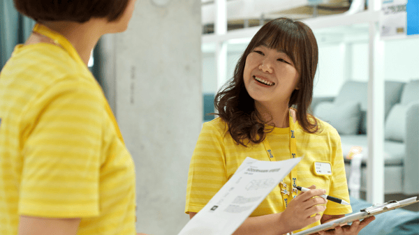 イケア店舗のショールームで、コワーカー同士がクリップボードの書類を確認しながら売場計画を話し合う場面。