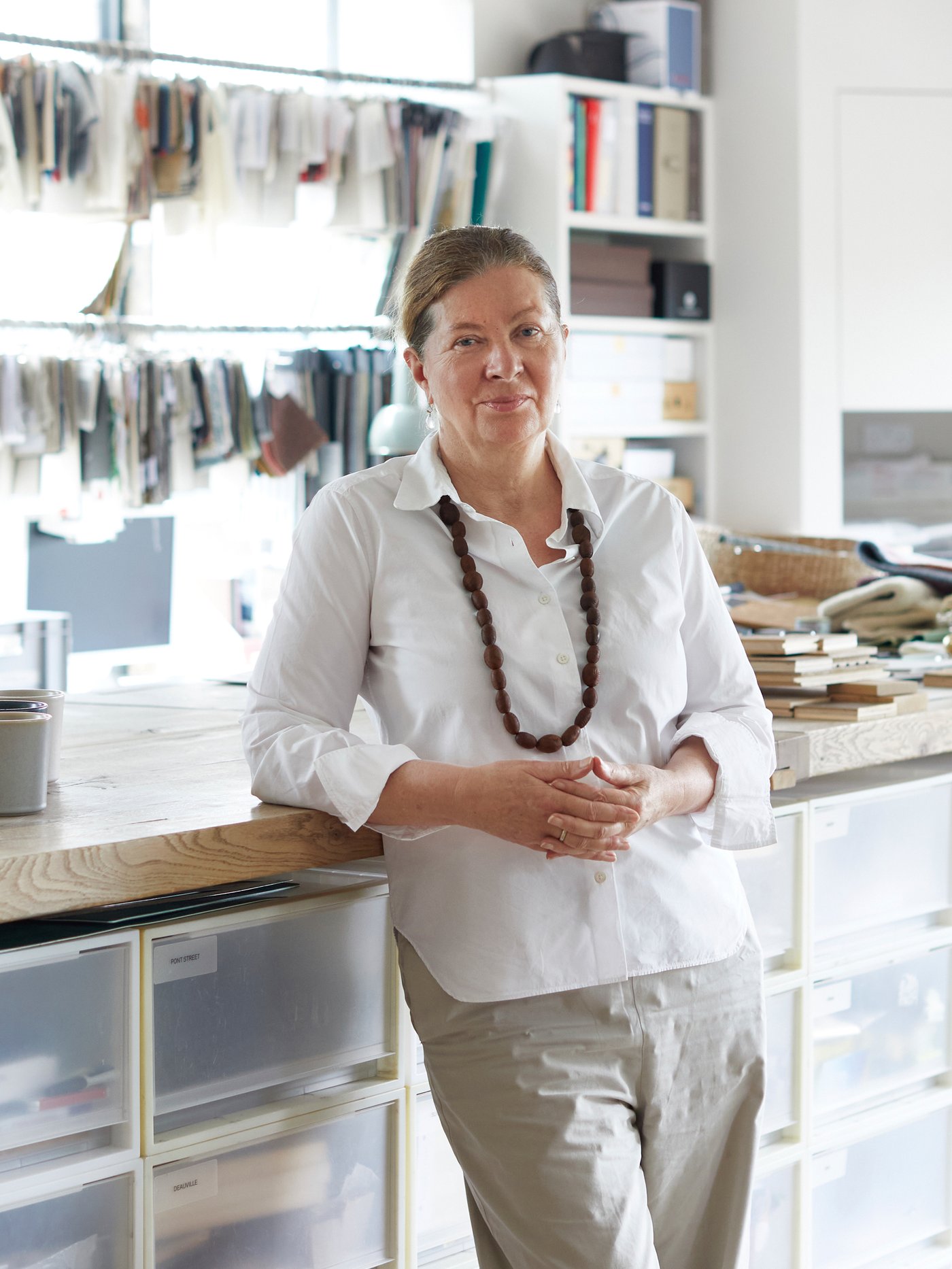 Ilse Crawford diseinatzailea, alkandora zuri batekin jantzita, laneko gainazal baten kontra makurtuta dago bere estudioan.