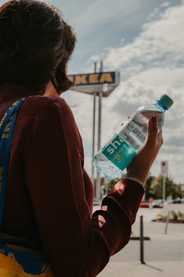Im Hintergrund ist das IKEA Logo zu sehen und im Vordergrund wird eine Wasserflasche von share gehalten.