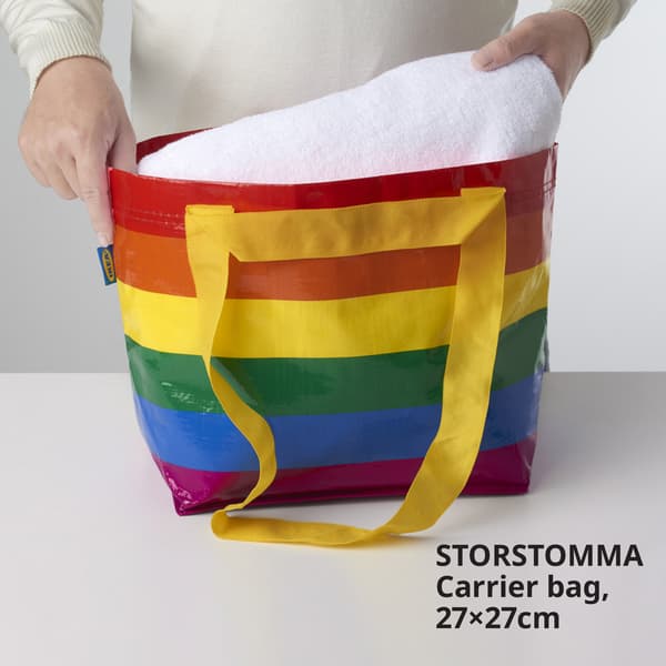 Image of a pair of hands putting a towel into a STORSTOMMA carrier bag, 27x27cm, with colourful strips.