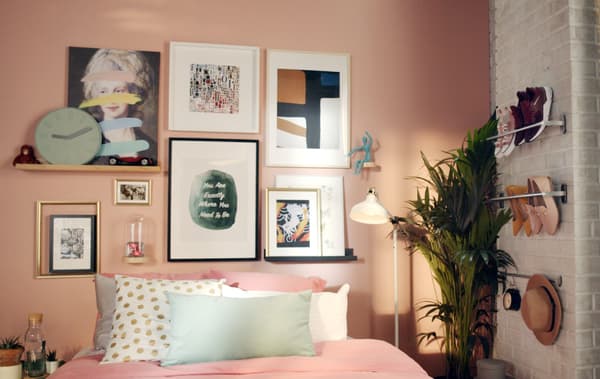 Imagem de uma cama encostada a uma parede, com bastantes almofadas. Na parede, em rosa, vê-se uma composição de molduras em diferentes cores, tamanhos e estilos.