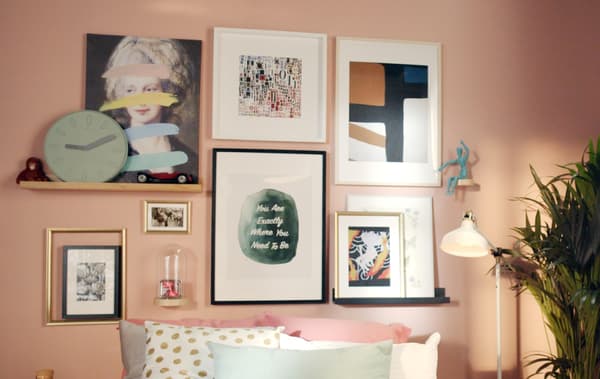 Imagem de uma cama encostada a uma parede, com bastantes almofadas. Na parede, em rosa, vê-se uma composição de molduras em diferentes cores, tamanhos e estilos.
