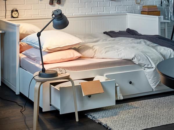 Imagen de una cama blanca desarmada, con cajones a medio abrir en la parte de abajo e iluminada por una lámpara de escritorio azul petróleo apoyada en un piso de madera usado a modo de velador. 