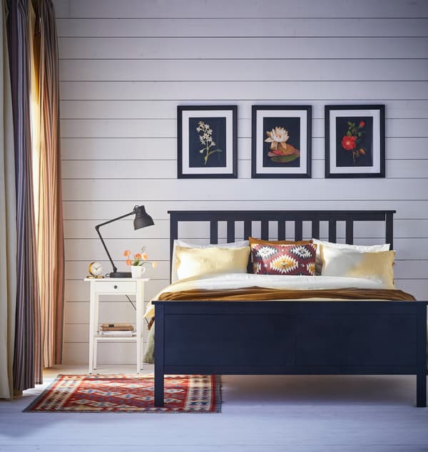 Imagen frontal de un dormitorio con una cama con respaldo, con tres cuadros sobre ella. Al costado izquierdo hay un velador con una lámpara de noche.