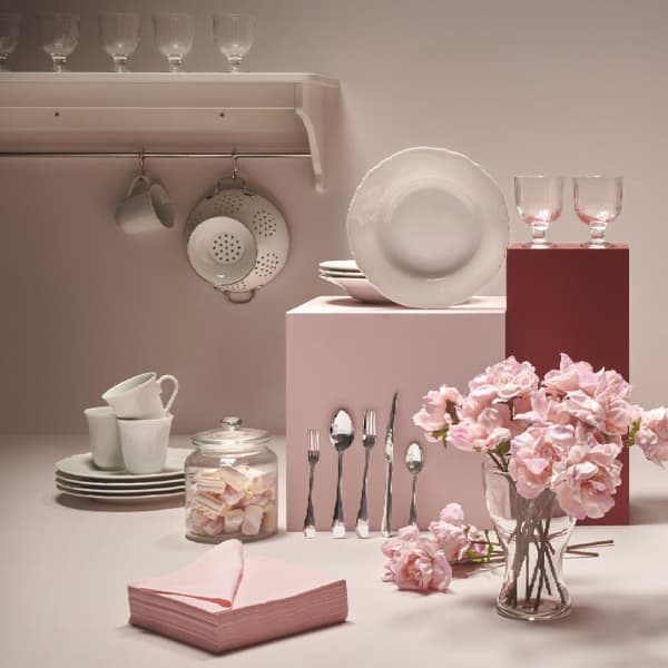 Imagen frontal de un set de vajilla UPPLAGA en un fondo beige y sobre una superficie rosado viejo, junto con un florero de rosas. 