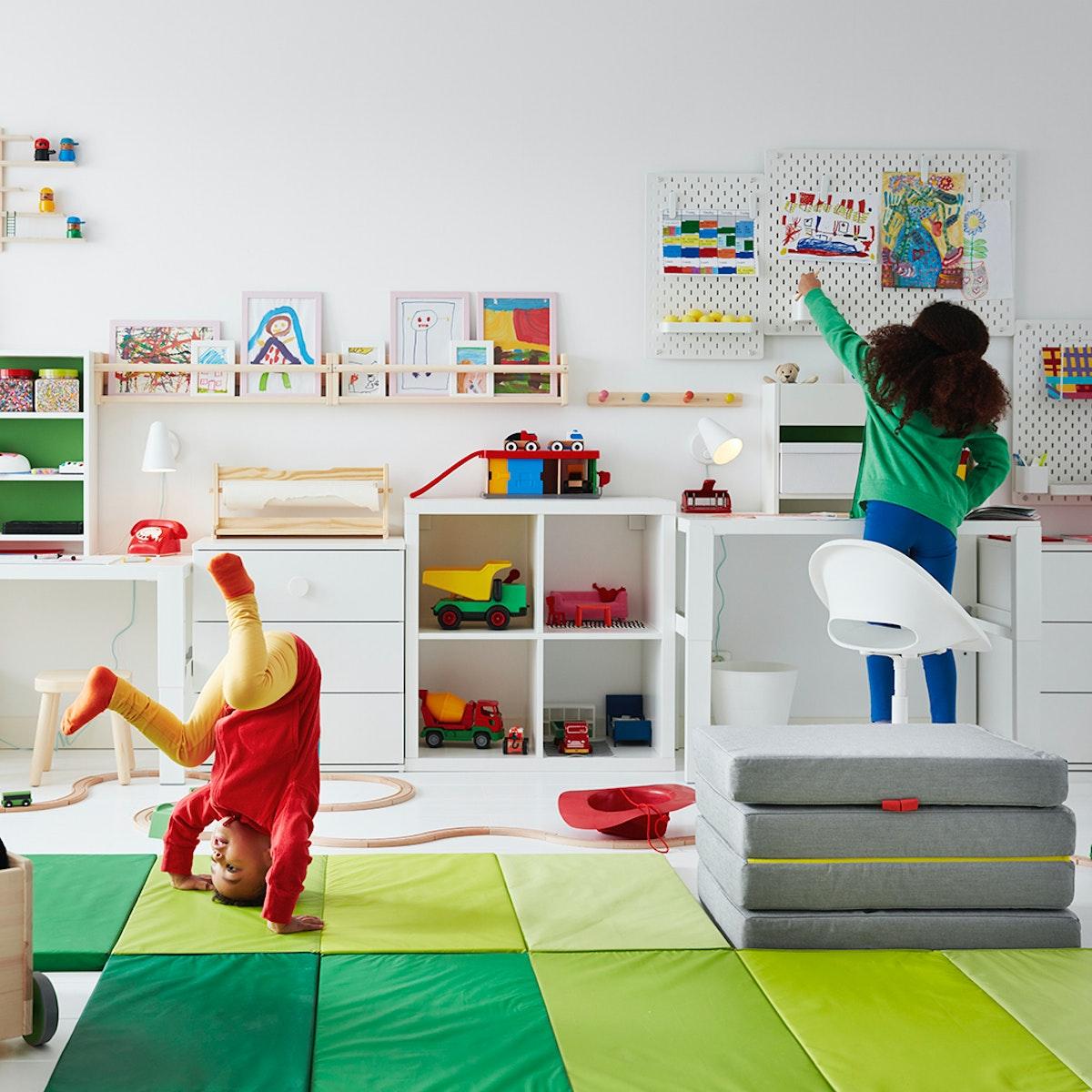 Imagen frontal de una pieza infantil con colchonetas PLUFSIG en el suelo y muebles blancos apoyados en el muro para organizar juguetes y útiles. Hay dos niños en la foto, uno juega en las colchonetas y la otra cuelga accesorios de un tablero mural. 
