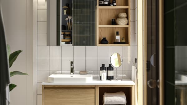 Imaginea arată o baie modernă cu mobilier din lemn deschis, chiuvetă albă, oglindă rotundă mică și rafturi deschise cu produse de îngrijire. Plante verzi în stânga și faianță albă pe pereți.