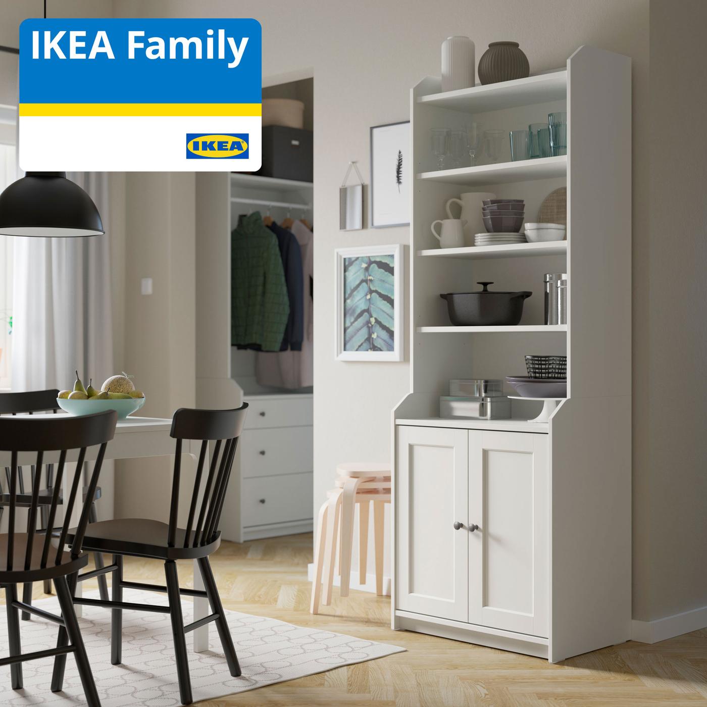 Imaginea arată o bucătărie modernă, cu un dulap alb deschis, plin cu vase și obiecte de bucătărie. În stânga sunt două scaune negre și o masă rotundă. În colțul din stânga sus este un logo IKEA Family.