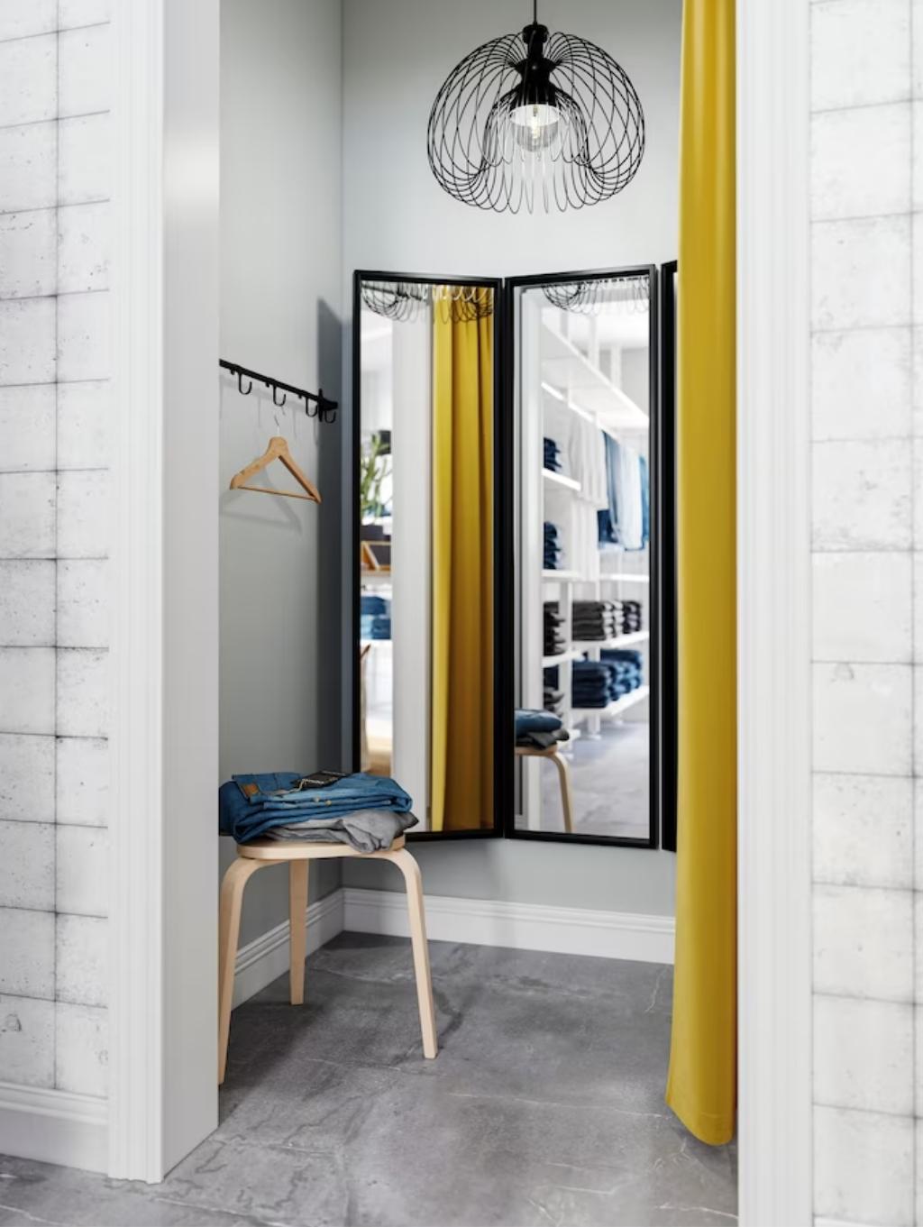 Imaginea arată o cabină de probă într-un magazin de haine. Cabina are un perete cu cârlige pentru umerașe, o oglindă triplă, un scaun din lemn pe care sunt așezate haine și o perdea galbenă. Deasupra este un corp de iluminat modern.