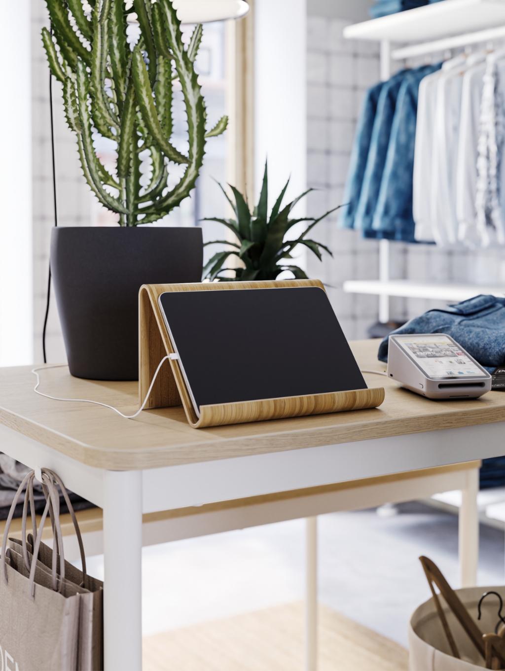 Imaginea arată o masă de lemn deschis la culoare pe care se află un suport de tabletă din lemn cu o tabletă neagră, un dispozitiv de plată electronic și două plante în ghivece. În fundal sunt haine agățate pe umerașe și o geantă maro sub masă.