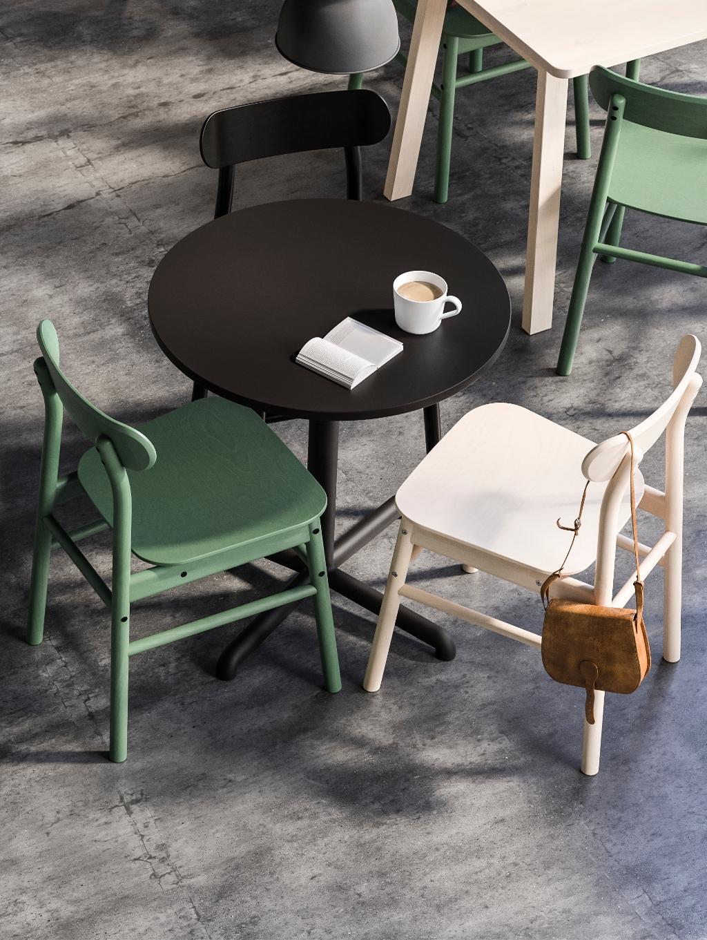 Imaginea arată o masă rotundă neagră cu două scaune verzi și un scaun alb. Pe masă se află o ceașcă de cafea și o carte deschisă. Un alt scaun alb are agățată de el o geantă maro. Podeaua este din beton gri.