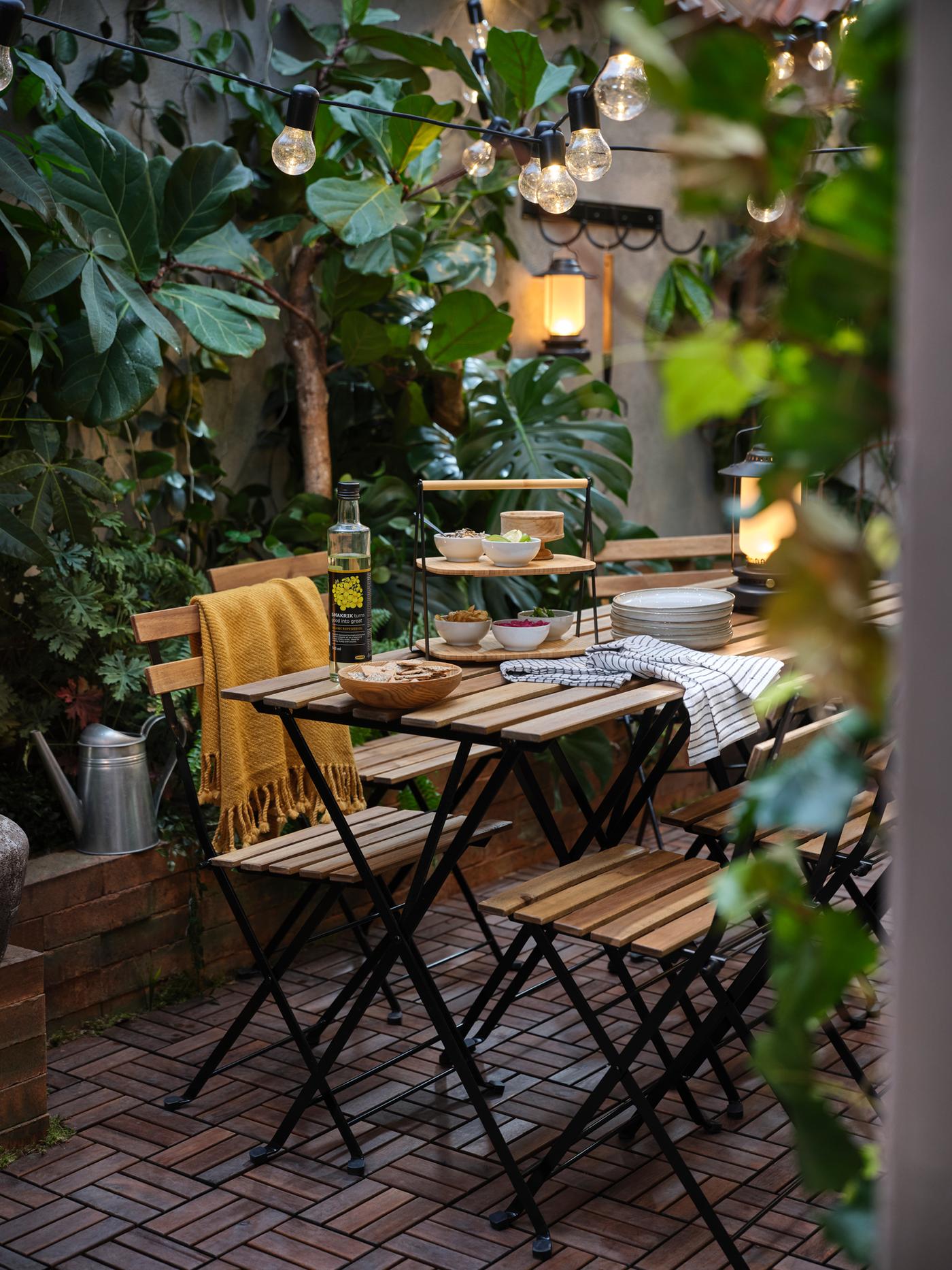 Imaginea arată o terasă exterioară cu o masă și scaune pliante din lemn, decorată cu plante verzi luxuriante. Pe masă sunt farfurii, pahare, o sticlă de apă și un bol cu mâncare. Deasupra mesei sunt agățate becuri decorative.