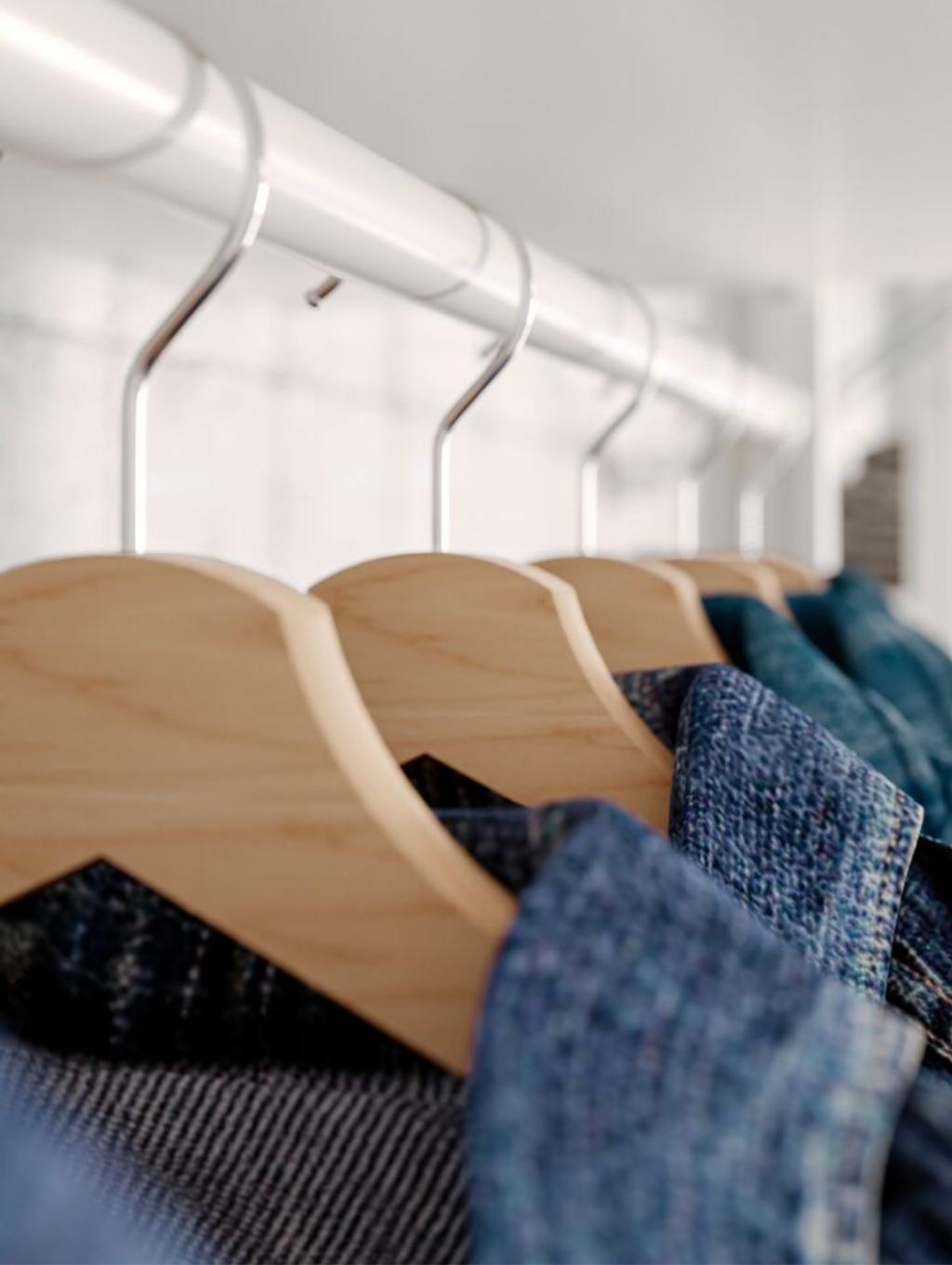 Imaginea arată patru umerașe din lemn pe un suport metalic alb, fiecare având agățate haine de culoare albastră. Umerașele sunt aranjate ordonat și sunt fotografiate dintr-un unghi lateral.