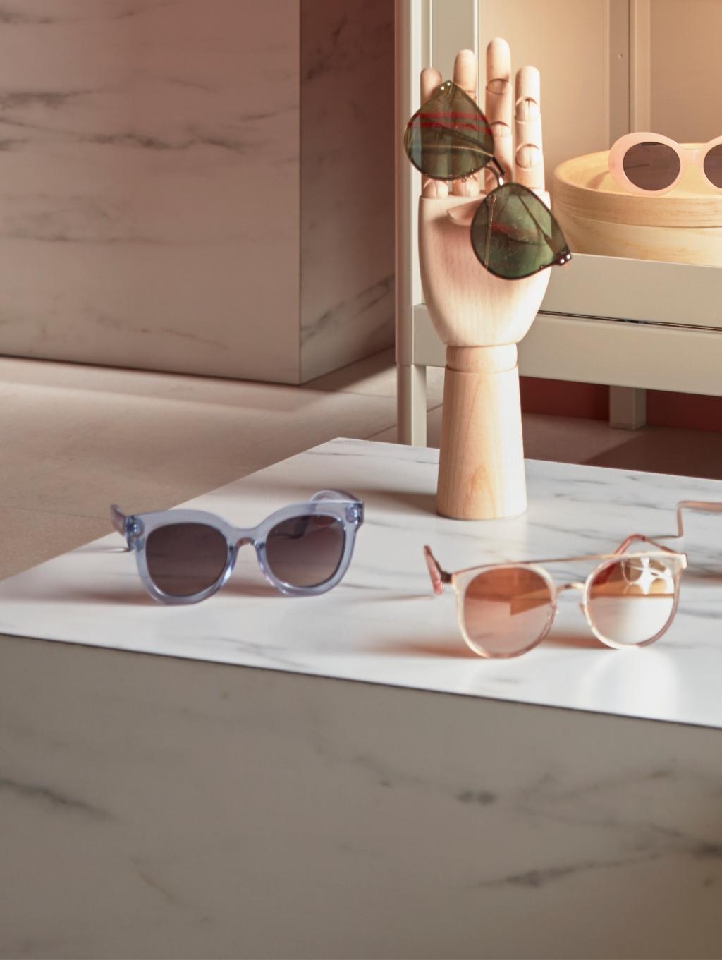 Imaginea arată trei perechi de ochelari de soare expuse pe o suprafață de marmură albă. O pereche este albastră, alta are lentile roz și rame aurii, iar a treia pereche este verde și este susținută de o mână manechin din lemn.