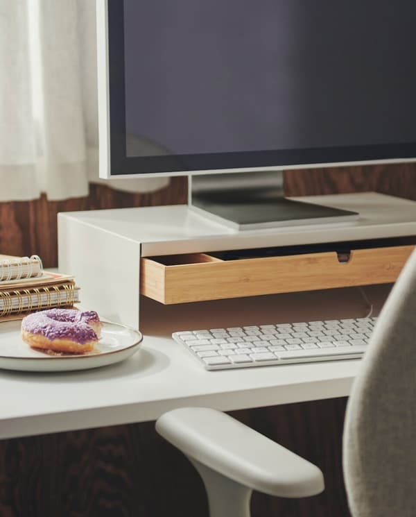 Imaginea arată un birou alb pe care se află o tastatură, un monitor mare, un sertar deschis din lemn, două caiete și o farfurie cu o gogoașă glazurată mov. În fundal se vede o perdea albă.