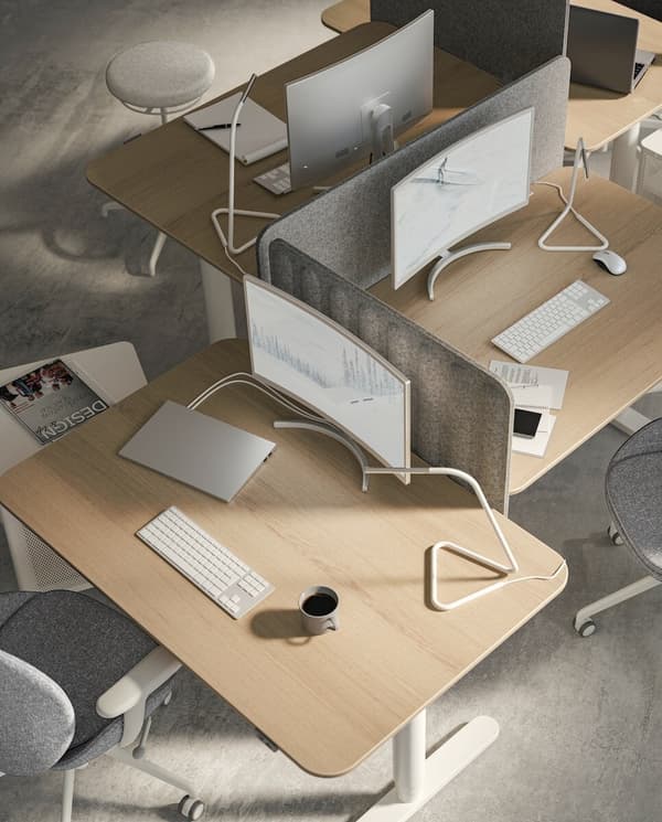 Imaginea arată un birou modern cu patru stații de lucru, fiecare echipată cu un monitor, tastatură și mouse. Pe birouri se află laptopuri, căști și o cană de cafea. Stațiile sunt separate de panouri gri pentru intimitate.